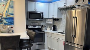 A modern kitchen with white cabinets, stainless steel appliances, and a dining area with a table and chairs. - Wave Action!Condo on the Beach w/Umbrella Service next to famous Wanna Wanna Bar (South Padre Island)