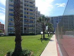 A modern multi-story building with balconies, a well-maintained lawn, and a pathway lined with trees and street lamps.