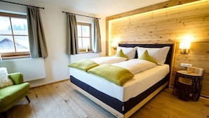 Quarto com cabeceira de madeira, duas camas com roupa de cama verde e branca, um banquinho de madeira e vista para o exterior pela janela.