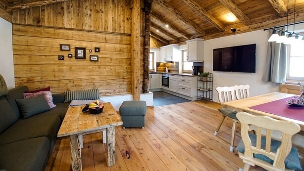 Interior acolhedor de uma cabana com teto de madeira, área de jantar com mesa e cadeiras, um sofá e uma cozinha ao fundo.