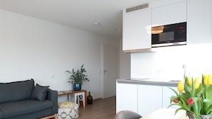 Una sala de estar moderna con un sofá, una mesita con un florero, y una cocina con armarios blancos y un microondas.