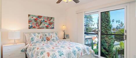 A bedroom with a bed, a nightstand, a lamp, a ceiling fan, a painting, and a sliding glass door leading to a balcony with a view of trees and buildings.