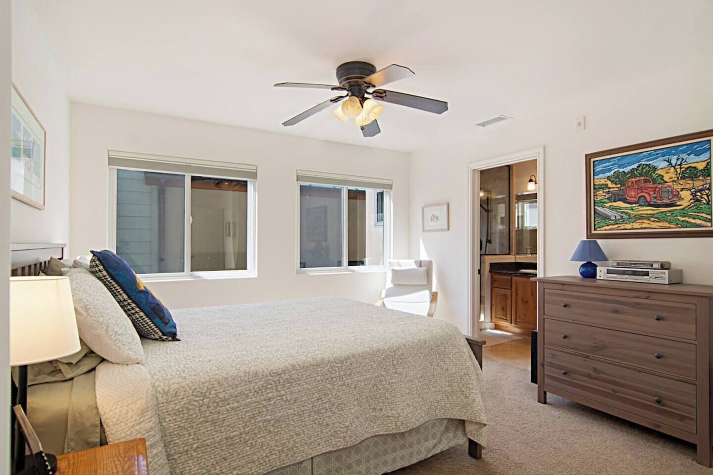 Ein Schlafzimmer mit Bett, Schminktisch, Schreibtisch, Stuhl, Deckenventilator und einem Bild an der Wand.