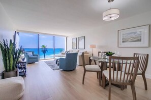 A spacious living area with a large window overlooking the ocean, a dining table with chairs, and a modern ceiling light.
