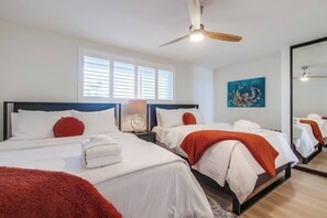 A hotel room with two beds, a ceiling fan, a large window with blinds, a painting on the wall, and a mirror reflecting the room.