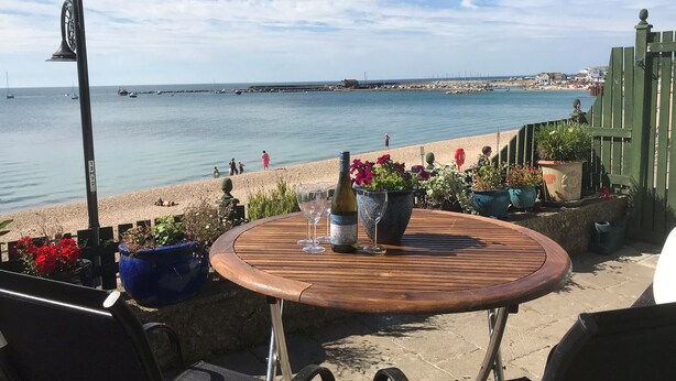 Una mesa de madera con una botella y un vaso, rodeada de plantas en macetas, con vistas a una playa donde se ven personas y barcos a lo lejos.