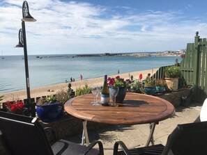A wooden table with a bottle and glass, surrounded by potted plants, overlooking a beach with people and boats in the distance. - Cottage on the Beach (Lyme Regis)