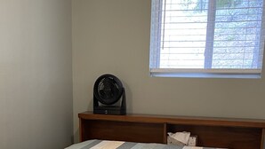 Una cama con colcha a rayas, cabecera de madera, ventana con persianas y un ventilador negro sobre un estante de madera.
