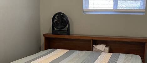 A bed with a striped bedspread, a wooden headboard, a window with blinds, and a black fan on a wooden shelf.