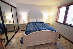 Een slaapkamer met een bed, een nachtkastje, een spiegel en een raam met jaloezieën.