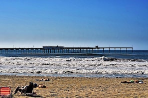 Une plage avec une jetée qui s’avance dans l’océan, une plage de sable et quelques personnes qui prennent un bain de soleil.