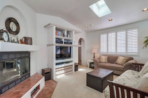 Un salon avec une cheminée, un téléviseur posé sur un meuble blanc, un canapé beige et une fenêtre avec des stores.
