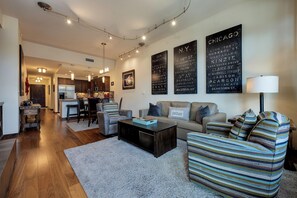 A modern living room with a sofa, armchairs, a coffee table, and a striped ottoman. There is a kitchen in the background with a dining area.
