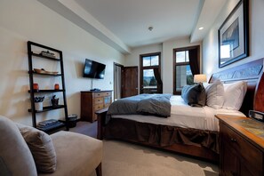 A hotel room with a bed, a TV, a wooden dresser, a bookshelf, and a window with a view.