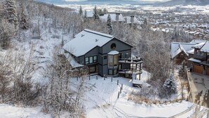 A modern house with a large balcony, surrounded by snow-covered trees and a mountainous landscape.