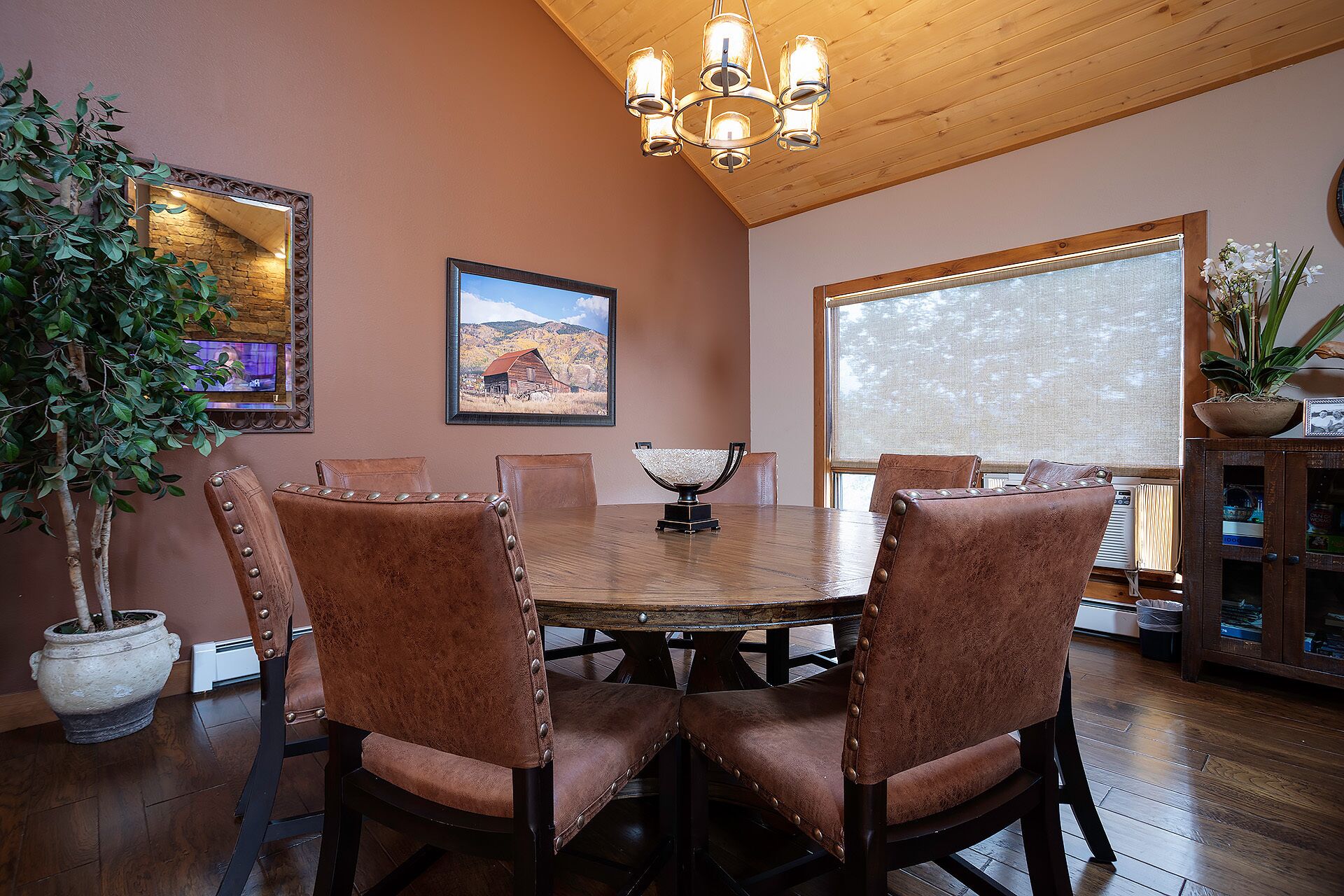 A dining area with a wooden table and chairs, a chandelier, a painting, and a large window.