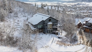 Una casa moderna con un balcón grande, rodeada de árboles cubiertos de nieve y un paisaje montañoso.