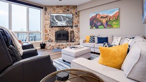 A living room with a stone fireplace, a large TV, and a sofa with colorful cushions. - SL300 | Modern Penthouse | Views | Pool | Hot Tub | 100 Yds to Lift (Steamboat Springs)