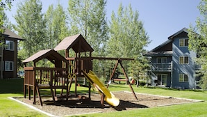 A playground with a slide and climbing structures, surrounded by a well-maintained lawn and trees.
