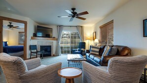 A living room with a fireplace, a ceiling fan, and a large window with blinds.