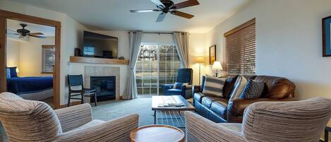 A living room with a fireplace, a ceiling fan, and a large window with blinds.