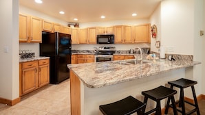 A kitchen with wooden cabinets, a black refrigerator, a microwave, an oven, a sink, and a granite countertop. There are two black barstools at the island.