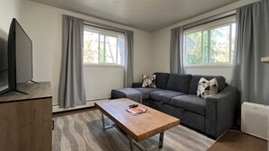 Una sala de estar moderna con un sofá seccional gris, una mesa de centro de madera, un televisor de pantalla plana y ventanas grandes con cortinas grises.