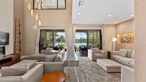 Un salon spacieux avec un grand canapé d’angle, une table basse et un téléviseur posé sur un meuble en bois. De grandes fenêtres sont agrémentées de rideaux, un ventilateur de plafond est installé et la pièce offre une vue sur l’extérieur.