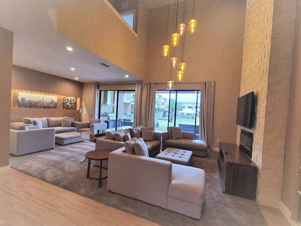 A spacious living room with a large sofa set, a coffee table, and a fireplace. There is a television mounted on the wall and a large window providing a view of the outside.