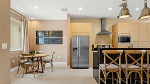 A modern kitchen with wooden cabinets, a stainless steel refrigerator, and a dining area with wooden chairs and a table. - 7648 Magic Village 8 guests 4 suites 8 min Disney  (Kissimmee)