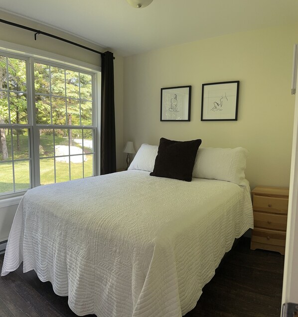 A bedroom with a large bed, two framed artworks on the wall, and a window overlooking a garden. - Sunlit cape on beautiful Bass Harbor close to Acadia National Park (Tremont)