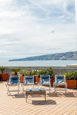 A terrace with sun loungers and potted plants, overlooking a coastal cityscape.