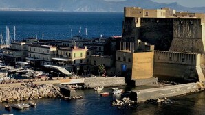 Un pueblo costero con una construcción fortificada, barcos y una vista despejada del mar y montañas distantes.