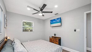 Een slaapkamer met een bed, een plafondventilator, een raam met zonwering, een aan de muur bevestigde televisie en een houten dressoir.