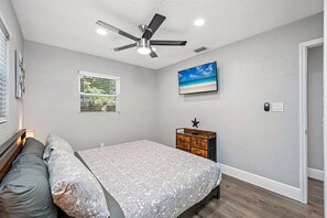 A bedroom with a bed, a ceiling fan, a window with blinds, a wall-mounted TV, and a wooden dresser.
