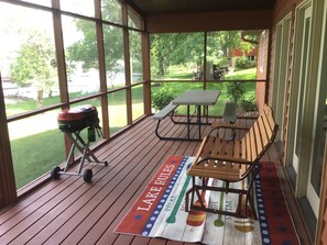 Une véranda abritée avec une table de pique-nique et un banc, un barbecue, et une vue sur une zone herbeuse et des arbres.
