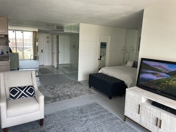 A modern hotel room with a bed, a TV, a seating area with a chair and ottoman, and a view of the outdoors.