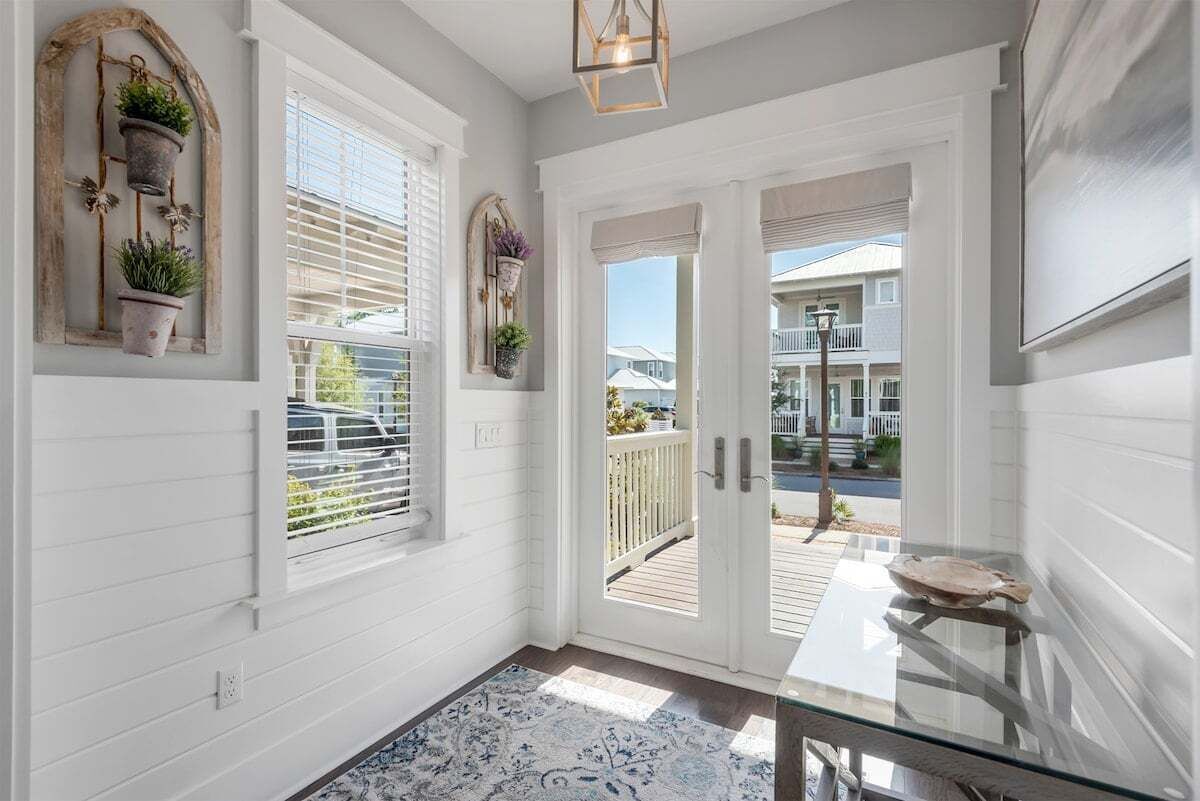 A hallway with white walls, a glass table, and a view of a deck and houses outside.