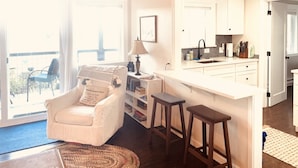 A bright living room with a white sofa, a coffee table, and a bookshelf. There is a kitchen area with white cabinets and a bar stool.