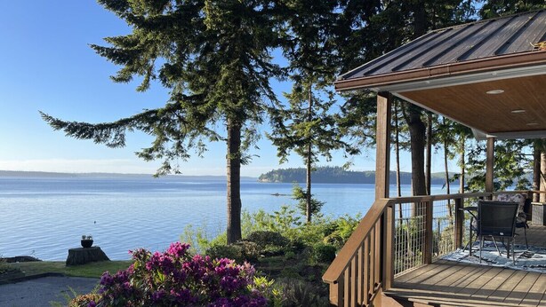 A wooden deck with outdoor furniture overlooking a lake, surrounded by trees and shrubs.