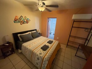 Quarto com cama, ventoinha no teto, mesa de cabeceira, mesa lateral e prateleira na parede.