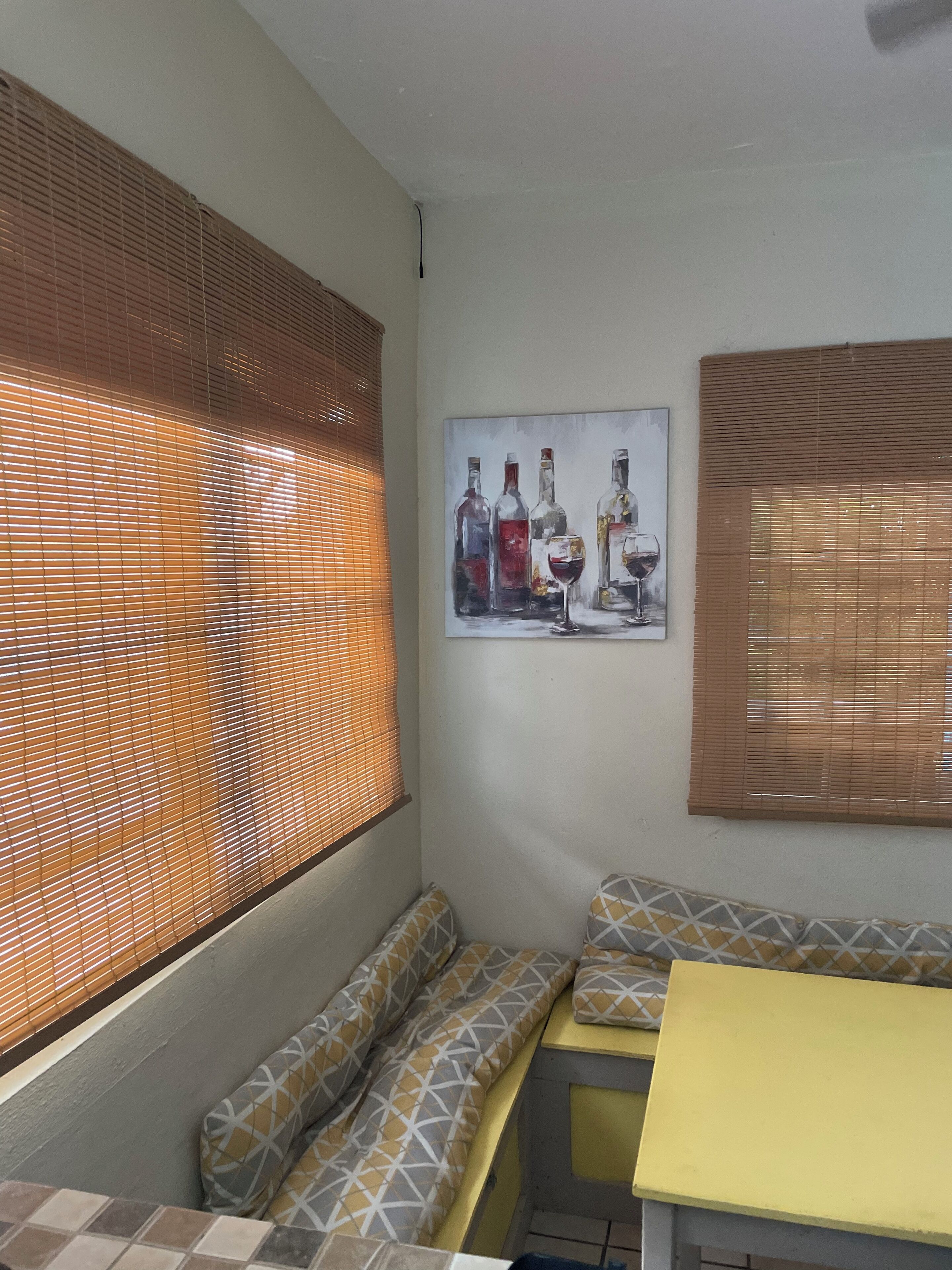 Une pièce d’angle avec un banc, une table jaune et un tableau représentant des bouteilles de vin accroché au mur.