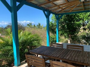 Um espaço ao ar livre coberto, com mesa e cadeiras de madeira, rodeado por vegetação exuberante e vista para a paisagem.
