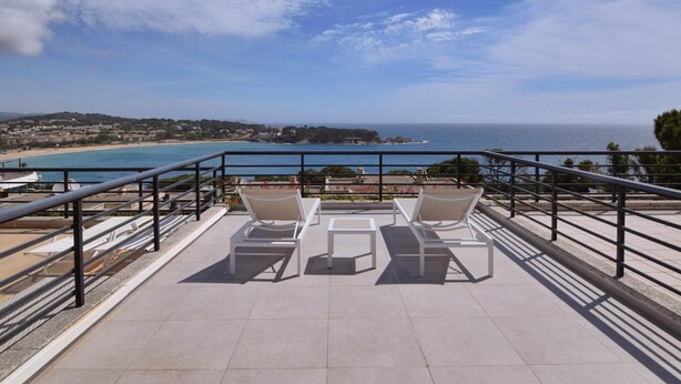 Eine Terrasse mit zwei weißen Liegestühlen und einem Tisch, mit Blick auf einen Strand und das Meer.