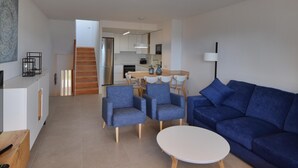 Ein modernes Wohnzimmer mit einem blauen Sofa, zwei blauen Sesseln und einem weißen Couchtisch. Im Hintergrund ist eine Küche mit einem Esstisch und Stühlen erkennbar.