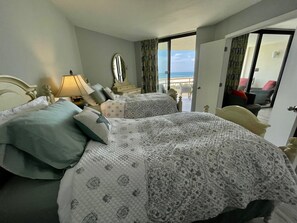 A hotel room with two beds, a nightstand, a lamp, a mirror, and a view of the beach through an open door.