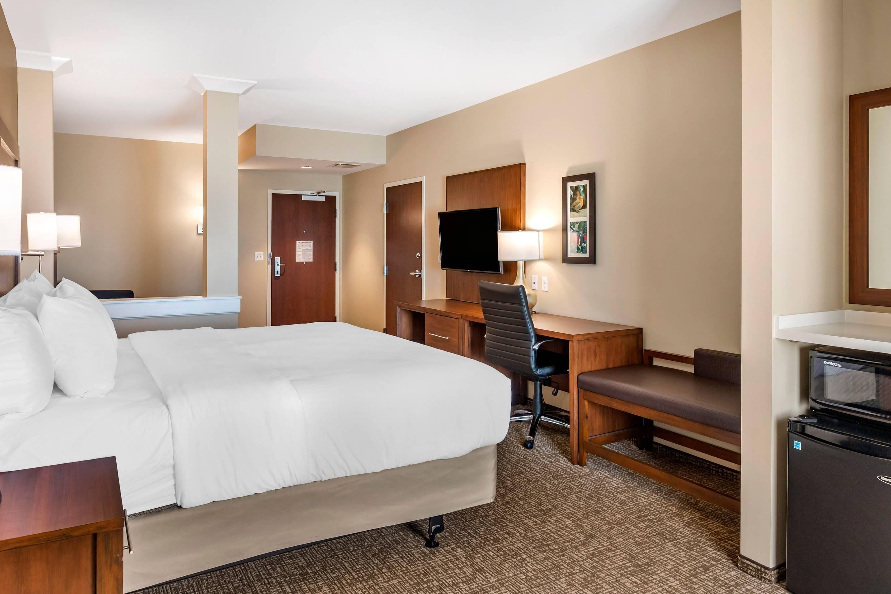 A hotel room with a large bed, a desk with a chair, a television, and a refrigerator.