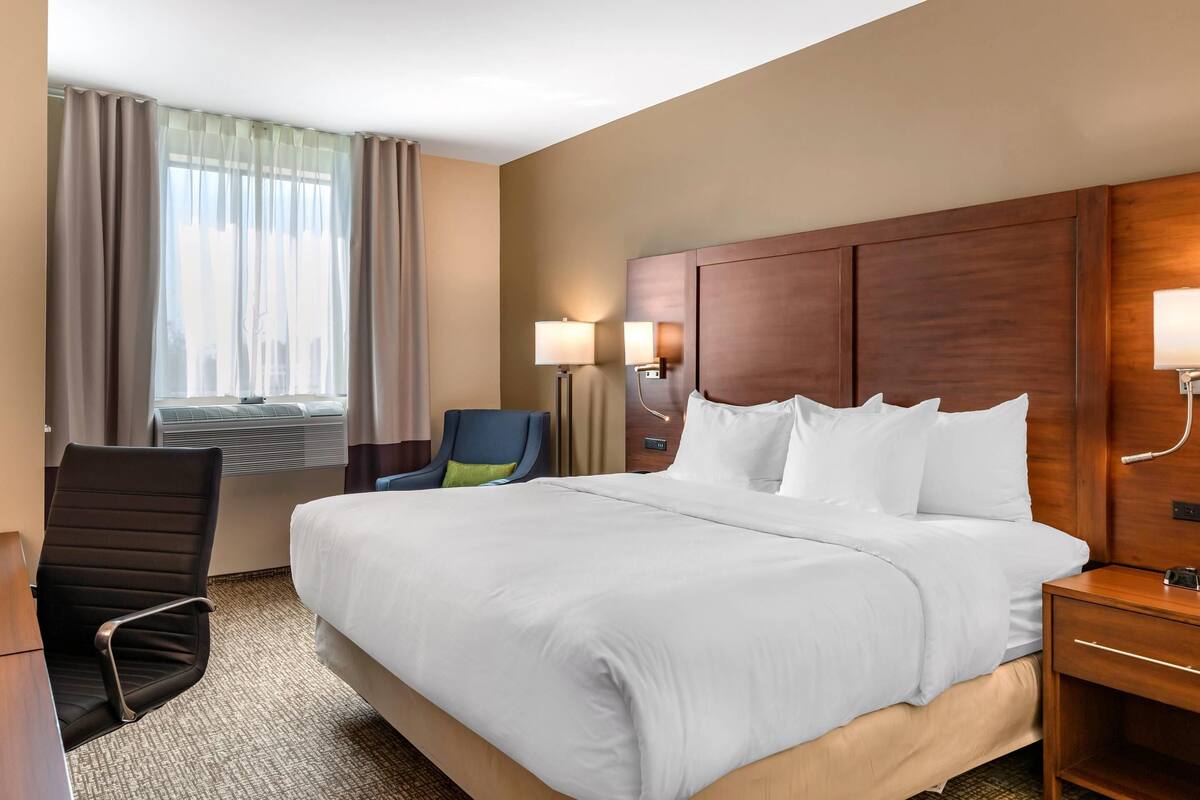 A hotel room with a large bed, a desk, a chair, a window with curtains, and wall-mounted lamps.