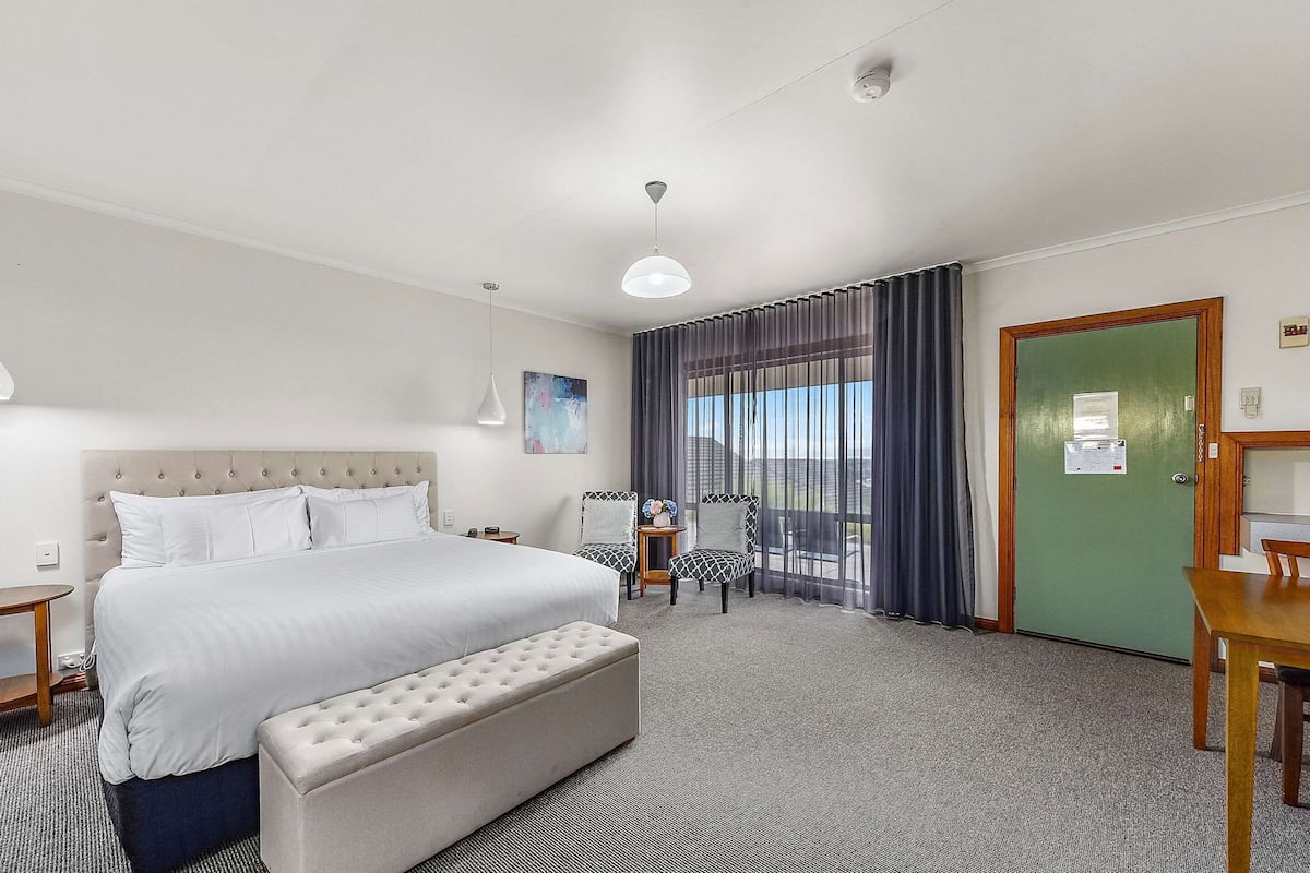 Ein Hotelzimmer mit Bett, einer Bank, einem Schreibtisch, einem Stuhl, einem Tisch, einem Fenster mit Vorhängen, einem Gemälde und einer grünen Tür.