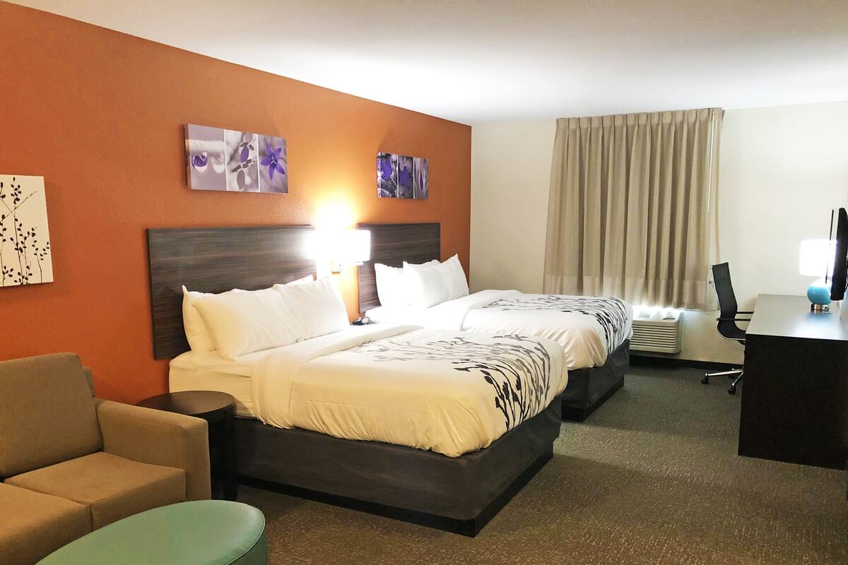 A hotel room with a double bed, a desk with a computer, a chair, a sofa, and a small ottoman.